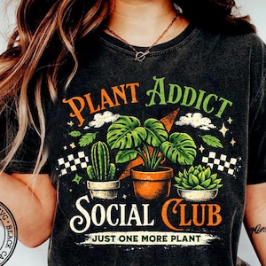 May include: Dark gray t-shirt with the words "PLANT ADDICT SOCIAL CLUB" and illustrations of plants. The shirt features a cactus, a monstera plant, and a succulent in pots. The text "JUST ONE MORE PLANT" is also included.