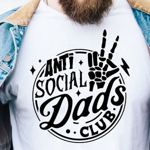 May include: A white t-shirt with a black graphic design that says "Anti Social Dad's Club" in a vintage style font. The graphic also includes a skeleton hand making a peace sign.