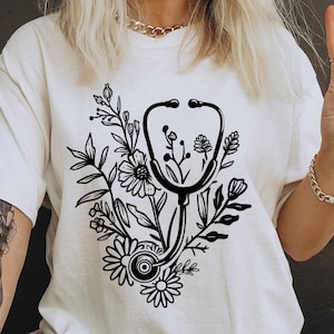 Op de afbeelding: Wit T-shirt met een zwart-wit grafisch ontwerp van een stethoscoop met bloemmotieven. De tekst "Black Cats" is zichtbaar in de rechterbenedenhoek van de afbeelding.