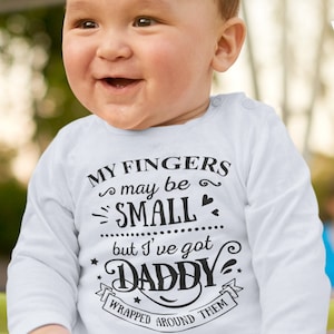 May include: A white long-sleeved onesie with black text that reads "My fingers may be small but I've got Daddy wrapped around them". The onesie is worn by a baby with a happy expression.