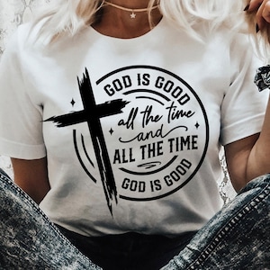 Puede incluir: Una camiseta blanca con un diseño gráfico negro que presenta una cruz y el texto "GOD IS GOOD all the time and ALL THE TIME GOD IS GOOD".