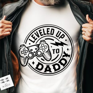 May include: White t-shirt with a black graphic featuring two game controllers and the text "LEVELED UP TO DADDY" in a circular design. The design includes a lightning bolt and star accents.