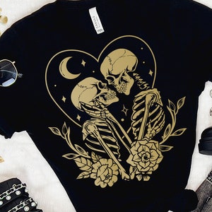 May include: Black t-shirt featuring a gold design of two skeletons embracing within a heart shape, adorned with stars and a crescent moon. The skeletons are holding flowers.
