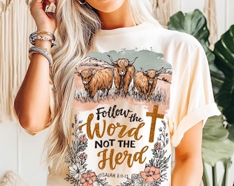 Può includere: T-shirt color crema con grafica di tre mucche delle Highlands in un campo, con la scritta "Follow the Word Not the Herd" e una croce. Elementi floreali e botanici circondano il testo. Isaia 8:11-13 è stampato sotto.