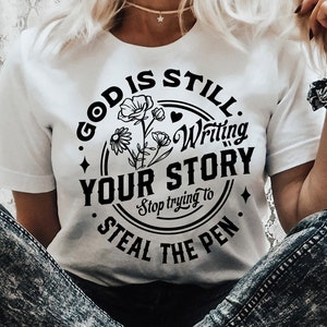 May include: A white t-shirt with a black circle design that says "God is still writing your story. Stop trying to steal the pen." The design also includes a floral illustration and a heart.