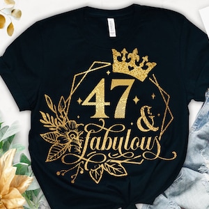 以下が含まれることがあります： 47の上に王冠が置かれた「47 & Fabulous」という金色のグリッターデザインが施された黒いTシャツ。デザインは幾何学的な形と花のアクセントで囲まれています。