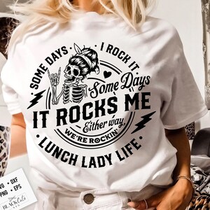 Może przedstawiać: Biała koszulka z czarno-białym wzorem graficznym przedstawiającym szkielet z rockandrollowym gestem. Tekst brzmi "Some days I rock it. Some days it rocks me. Either way, we're rockin'. Lunch Lady Life."
