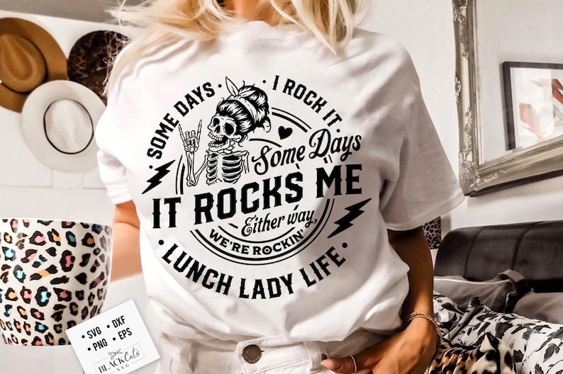 Może przedstawiać: Biała koszulka z czarno-białym wzorem graficznym przedstawiającym szkielet z rockandrollowym gestem. Tekst brzmi "Some days I rock it. Some days it rocks me. Either way, we're rockin'. Lunch Lady Life."