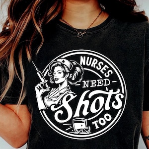Puede incluir: Diseño gráfico en blanco y negro sobre una camiseta negra con una enfermera de dibujos animados con una jeringa y el texto "Nurses Need Shots Too".