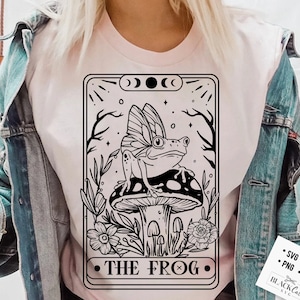 Op de afbeelding: Een zwart-witte illustratie van een kikker met vleugels die op een paddenstoel zit. De illustratie is in de stijl van een tarotkaart en heeft de tekst "THE FROG" onderaan.