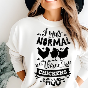 May include: White sweatshirt with black text that reads "I was normal three chickens ago". The text is surrounded by decorative elements, including hearts and leaves. Three black silhouettes of chickens are shown below the text.