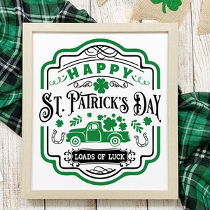 以下が含まれることがあります： 白地に緑と黒の看板。看板には「Happy St. Patrick's Day」と、クローバーでいっぱいのトラックと「Loads of Luck」の文字が書かれている。