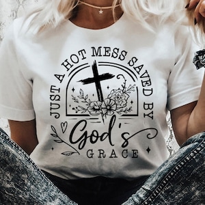 May include: White t-shirt with a black graphic design. The design includes a cross, floral elements, and the text "JUST A HOT MESS SAVED BY GOD'S GRACE". The shirt also has the text "SVG, DXF, PNG, EPS" and the BlackCats SVG logo.