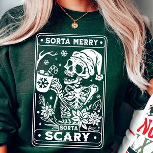 May include: A green sweatshirt with a white graphic of a skeleton wearing a Santa hat and holding a mug of hot cocoa. The graphic is surrounded by a white border and includes the text "Sorta Merry" and "Sorta Scary".