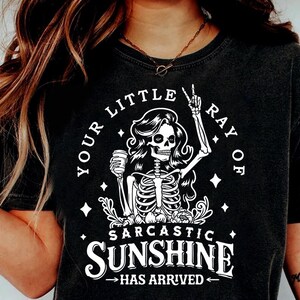 May include: Black t-shirt with a white graphic of a skeleton holding a coffee cup and giving a peace sign. Text reads "YOUR LITTLE RAY OF SARCASTIC SUNSHINE HAS ARRIVED". The shirt is a dark charcoal color.
