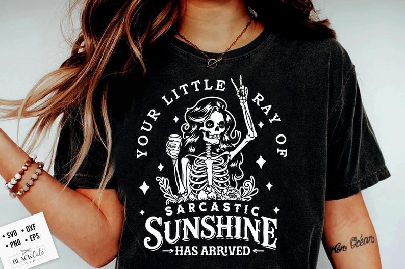 May include: Black t-shirt with a white graphic of a skeleton holding a coffee cup and giving a peace sign. Text reads "YOUR LITTLE RAY OF SARCASTIC SUNSHINE HAS ARRIVED". The shirt is a dark charcoal color.
