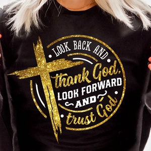 Może przedstawiać: Czarna bluza z złotym brokatowym krzyżem i napisem "LOOK BACK AND thank God, LOOK FORWARD AND trust God." Projekt jest okrągły z nakładką w kształcie krzyża.