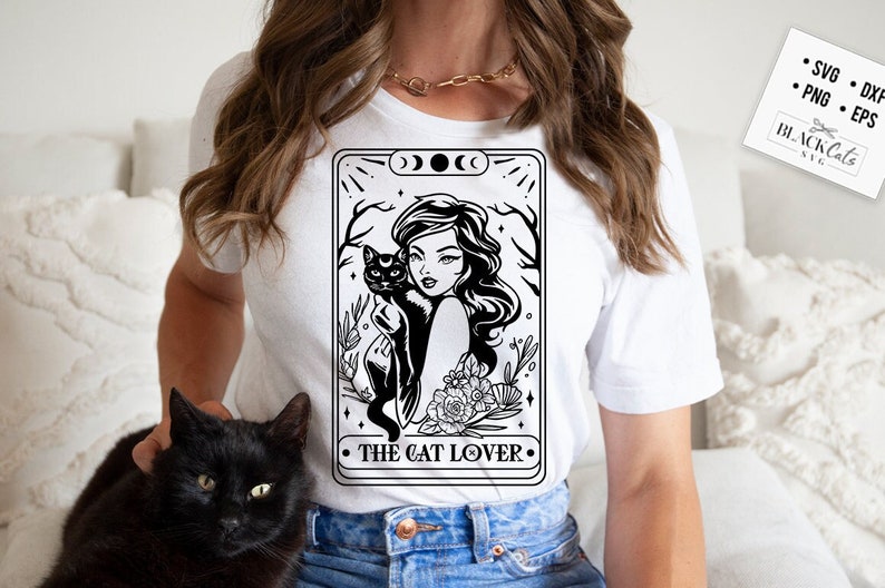 May include: White t-shirt featuring a black and white graphic of a woman holding a black cat, with the text "THE CAT LOVER". The design includes floral and celestial elements. A black cat is also visible in the image.