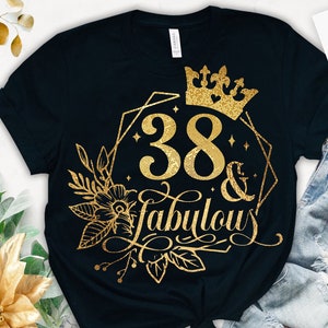 Może przedstawiać: Czarny t-shirt z złotym brokatowym wzorem, na którym widnieje napis "38 & fabulous" z koroną i kwiatowymi akcentami.