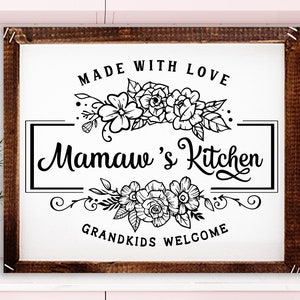 May include: A wooden framed sign with the text "MADE WITH LOVE" and "Mamaw's Kitchen" in a decorative font. Floral designs are above and below the text. The words "GRANDKIDS WELCOME" are at the bottom. The sign is white with black lettering.