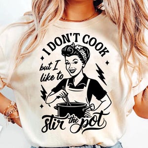 May include: Cream-colored t-shirt with a black graphic of a woman stirring a pot, with the text "I DON'T COOK but I like to Stir the pot". The design includes lightning bolts and stars, a retro kitchen theme.
