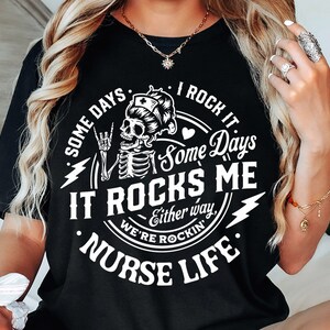 Peut inclure: T-shirt noir avec un motif blanc repr&eacute;sentant un squelette portant une coiffe d'infirmi&egrave;re, faisant un geste de rock and roll. Le texte dit "Some days I rock it, Some days it rocks me, Either way, we're rockin' Nurse Life."