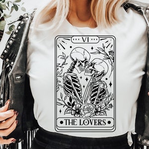 Puede incluir: Una ilustraci&oacute;n en blanco y negro de la carta del tarot de Los Enamorados que presenta dos esqueletos abraz&aacute;ndose. La carta est&aacute; enmarcada con un borde negro y el texto "VI" y "THE LOVERS" est&aacute; impreso en negro.