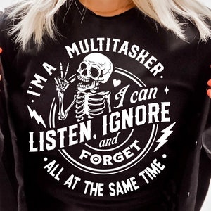 Puede incluir: Camiseta negra con un gráfico circular blanco que presenta un esqueleto con un signo de paz, un corazón y el texto "Soy un multitarea. Puedo escuchar, ignorar y olvidar todo al mismo tiempo."