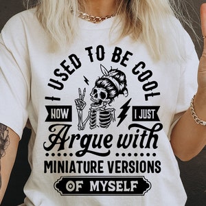 May include: A white t-shirt with a black graphic design that says "I used to be cool now I just argue with miniature versions of myself". The graphic includes a skeleton with a peace sign and lightning bolt.