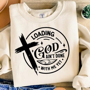 May include: A white sweatshirt with a black graphic design that says "Loading... God ain't done with me yet." The graphic also includes a cross.