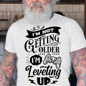 Puede incluir: Una camiseta blanca con texto negro que dice "No estoy envejeciendo, estoy subiendo de nivel" con una ilustración en blanco y negro de un mando de videojuego.
