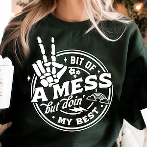 May include: A green sweatshirt with a white graphic design that says "Bit of a Mess but doin' my best". The graphic features a skeleton hand making a peace sign.