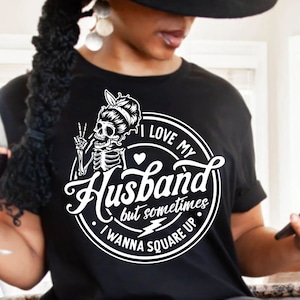 May include: Black t-shirt with a white circle graphic featuring a skeleton with a peace sign, a heart, and the text "I love my Husband but sometimes I wanna square up".