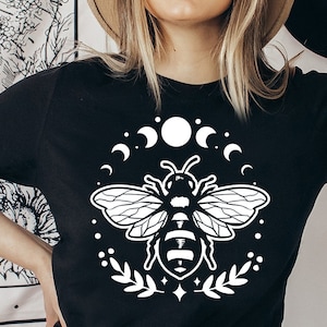 May include: Black t-shirt featuring a white graphic of a bee surrounded by crescent moons, dots, and leaves. The bee design is centered on the shirt. The shirt also has a small white text that says "Black Cats SVG".