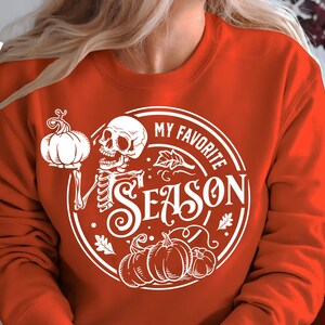 May include: Orange sweatshirt with a white graphic that says "MY FAVORITE SEASON" with a skeleton holding a pumpkin. The design also includes pumpkins and leaves. The sweatshirt has the words "SVG, DXF, PNG, EPS" and "BLACK CATS SVG" in the upper left corner.