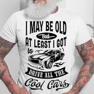 May include: A white t-shirt with a black graphic design that says "I may be old but at least I got to drive all the cool cars". The graphic also includes a black and white illustration of a classic car.