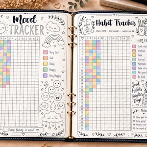 May include: An open ring-bound planner featuring a Mood Tracker on the left page and a Habit Tracker on the right. Both pages include grids with pastel-colored squares, black ink illustrations of clouds, rainbows, and faces, and text labels.