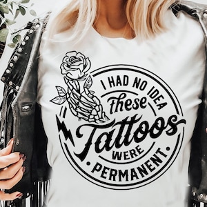 May include: White t-shirt with a black graphic featuring a skeleton hand holding a rose and the text "I had no idea these tattoos were permanent." The shirt is worn with a black leather jacket.