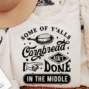 May include: A white sweatshirt with a black graphic design that says "Some of y'alls cornbread ain't done in the middle". The graphic also includes a skillet with cornbread and a pointing hand.