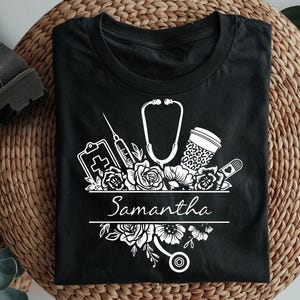 May include: Black t-shirt with a white design of a stethoscope, syringe, bandage, coffee cup, and flowers. The text "Samantha" is written in a banner below the design.