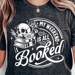May include: A white graphic design on a dark grey t-shirt with a skull, flowers, and books. The text reads "My Weekend Is All Booked".