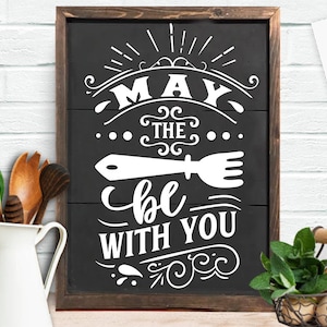 May include: A black wooden sign with white text that reads "May the fork be with you." The sign has a rustic, farmhouse style and is framed in dark brown wood. The image also includes a white pitcher, wooden utensils, and a basket of herbs.