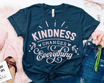 Puede incluir: Una camiseta azul oscuro con un gráfico rosa que dice "Kindness Changes Everything". El gráfico está en una fuente estilizada con un corazón y acentos florales.