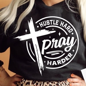 以下が含まれることがあります： 白い十字架と「Hustle Hard Pray Harder」というテキストが描かれた白いグラフィックデザインが施された黒いスウェットシャツ。