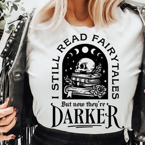 Puede incluir: Una camiseta blanca con un diseño gráfico negro que presenta una calavera, libros y el texto "I still read fairytales but now they're darker".