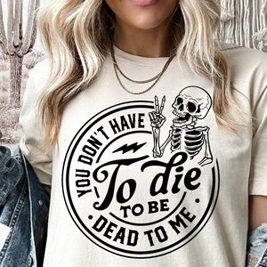 Può includere: Una maglietta bianca con un grafico circolare nero che presenta uno scheletro con un segno di pace, il testo "You don't have to die to be dead to me" e un fulmine.