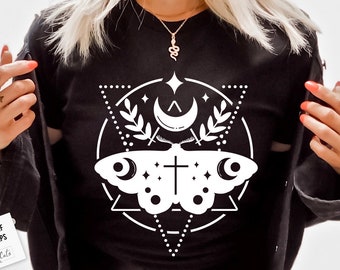 May include: A black t-shirt with a white graphic design of a moth with a cross in the center, surrounded by a crescent moon, stars, and a triangle. The design is in a geometric style.