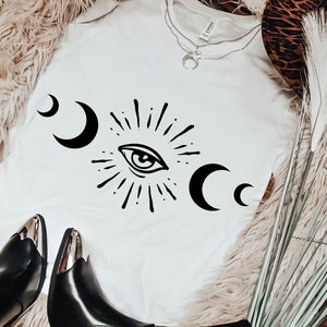 May include: A white t-shirt with a black and white design of an eye with a sunburst and crescent moons. The shirt is laying on a fuzzy brown surface with a brown and white animal print surface and a bouquet of dried grass.
