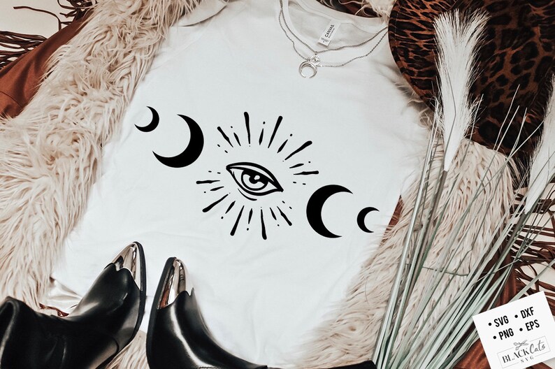 May include: A white t-shirt with a black and white design of an eye with a sunburst and crescent moons. The shirt is laying on a fuzzy brown surface with a brown and white animal print surface and a bouquet of dried grass.