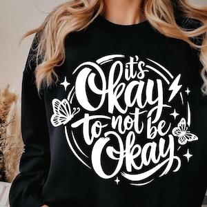 May include: Black sweatshirt with the white text "It's Okay to not be Okay" in a circular design with butterfly and star graphics. The sweatshirt is a crew neck style.